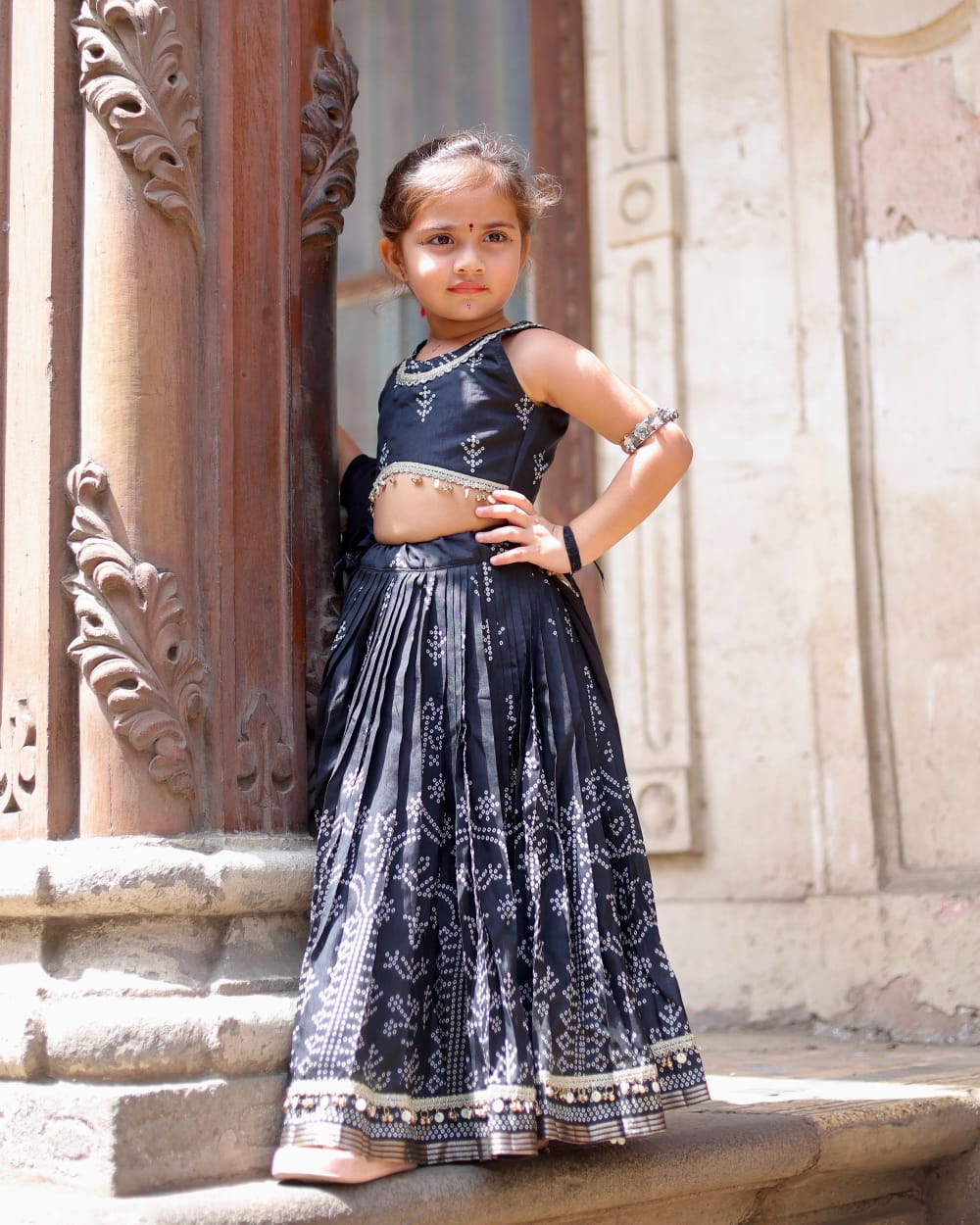 A girl in an indigo bandhani lehenga standing on a stone step beside a carved pillar