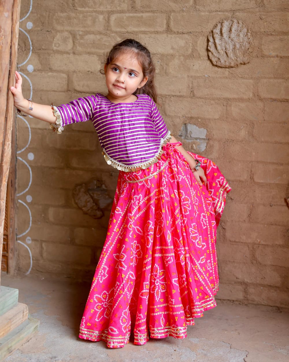 A girl in a rani pink bandhani ghagra holding the skirt out beside a wooden post