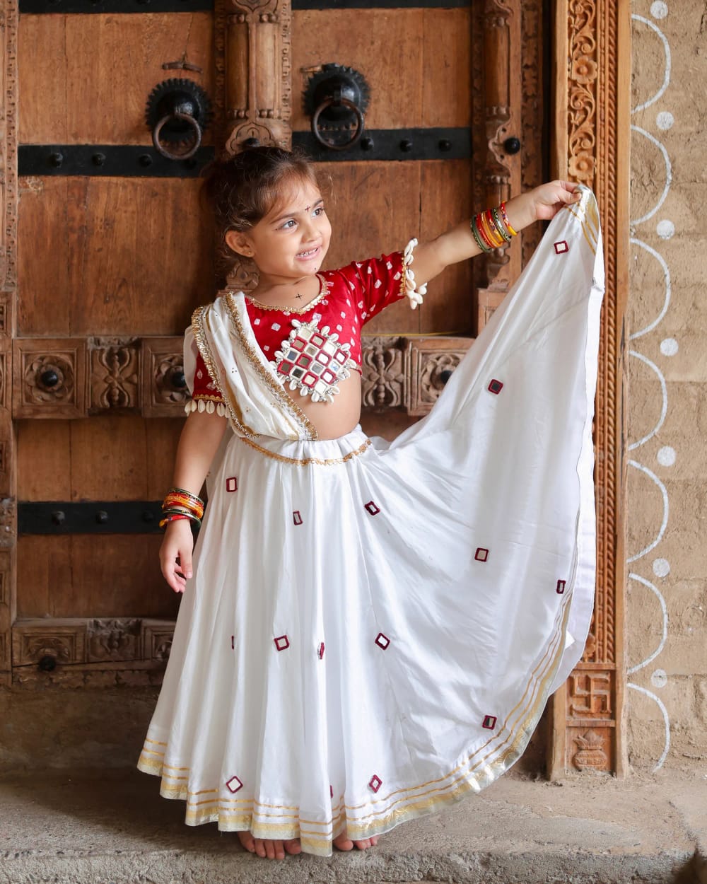 A girl holding the white dupatta open in front of carved wooden doors